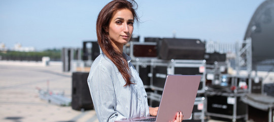 Installation of stage equipment and preparing for a live concert open air. Event manager portrait. Summer music city festival. Young serious woman stand and work with her laptop near the stage.